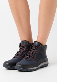 Zapatillas altas de color azul oscuro con acentos en naranja, materiales de cuero y textil, cuello acolchado y suela de goma texturizada.
