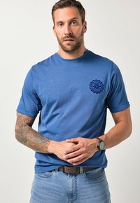 Blue cotton t-shirt with short sleeves and a round neckline. Features a dark blue logo on the left chest. Smooth texture, casual fit.
