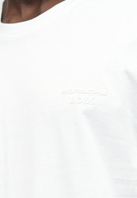 Κοντινό πλάνο σε λευκό μπλουζάκι με ανάγλυφο λογότυπο "Porsche Boss" στο αριστερό στήθος, που φορεί ένα άτομο με σκουρόχρωμο δέρμα.