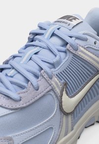 Ανοιχτό μπλε αθλητικό παπούτσι Nike με λευκό λογότυπο Swoosh, υφασματινές επιφάνειες με υφή και ετικέτα "P H O M E R O 5" στη γλώσσα, κοντινή εικόνα δετών και επάνω μέρους.