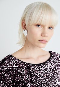 Strieborné náušnice v tvare srdca, lesklý povrch, nosené modelkou s krátkymi blond vlasmi. Vrchná časť má čierny výstrih a fialové flitrové zdobenie.