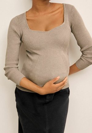 Kobieta w ciąży w beżowym prążkowanym swetrze i czarnej spódnicy, delikatnie trzymająca brzuch jedną ręką.