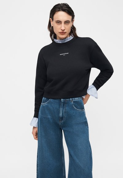 Femme portant un sweat-shirt noir Maison Kitsuné par-dessus un chemisier bleu clair à volants et un jean bleu taille haute à jambes larges, debout avec une main sur la hanche.