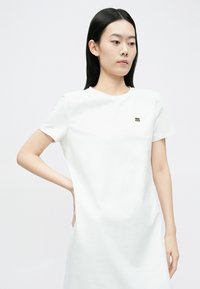 Valge puuvillane t-särgi kleit ümarate kaelusega, lühikeste varrukatega ja väike must logodetail rinnal. Sile tekstuur.