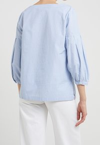 Chemisier rayé bleu et blanc avec manches bouffantes, col rond et fentes latérales. Fabriqué en tissu léger au toucher doux.