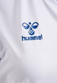 Vit sportskjorta med blå Hummel-logotyp, med en slät textur och atletisk design. Logotypen inkluderar en stiliserad bi och geometriska mönster.