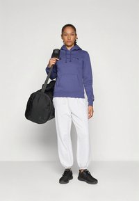 Sweatshirt roxo com capuz e logótipo bordado, calças de treino cinzentas e ténis pretos. O modelo segura uma mala desportiva preta.