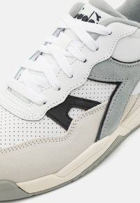 Witte sportieve schoen van geperforeerd leer, met grijze suède accenten, zwart logo-detail en platte witte veters. Rond neus en gevoerde zool.