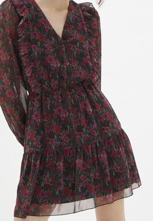 Femme portant une robe sombre transparente à motif floral, manches longues, détails à volants et jupe à étages sur un fond clair uni.
