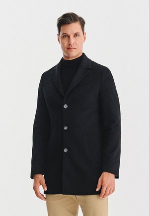 Czarny wełniany płaszcz z klapami, z czterema dużymi guzikami i bocznymi kieszeniami, noszony na czarnej golfie. Tkanina jest gładka.