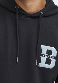 Sudadera negra con bolsillo frontal y cuerdas ajustables. Presenta una "B" bordada en azul claro y "BLEND" en el pecho. Textura suave.
