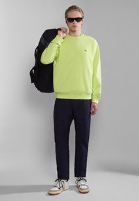 Sudadera verde neón con cuello redondo, pantalones cargo azul marino, zapatillas blancas con detalles en azul marino y bolsa de deporte negra. Las gafas de sol completan el look.