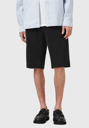 Pantaloni neri realizzati in un tessuto liscio, lunghezza al ginocchio, con una vestibilità comoda. Indossati con una camicia a righe blu chiaro e scarpe nere.