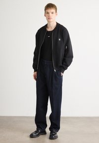 Ung mand står indendørs iført en sort zip-up bomberjakke, sort T-shirt, løse mørkeblå jeans og sorte lædersko.