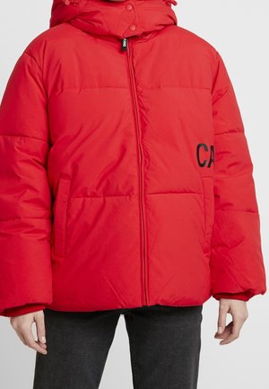 Červená péřová bunda se zipem vpředu, bočními kapsami, kapucí a částečným černým nápisem na pravé straně, nošená s černými kalhotami.