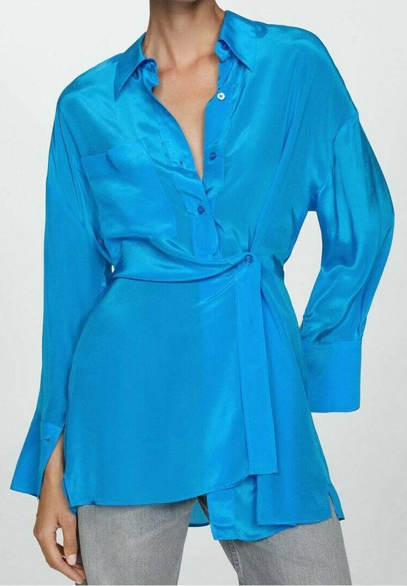 Femme portant un chemisier cache-cœur bleu vif en satin à manches longues avec des boutons à l'avant, des poignets surdimensionnés, et un pantalon gris.