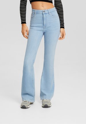 Jeans de tiro alto de mezclilla en azul claro, con diseño acampanado, que cuentan con cinco bolsillos y un cierre frontal con botón. Combinados con zapatillas grises.