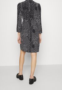 Svart klänning med varierande vita prickmönster, långa ärmar, hög krage och en lätt utsvängd fåll, tillsammans med svarta skor.