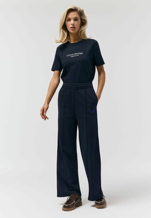 Blondynka stoi, ubrana w czarną koszulkę z napisem „Diverse Heritage”, luźne czarne spodnie z elastycznym pasem oraz brązowe buty sznurowane.