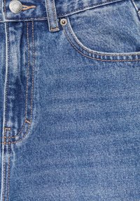 Gros plan sur un jean en denim bleu montrant la poche avant, la passants de ceinture, les coutures orange et un bouton métallique avec texte de la marque.
