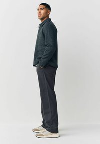 Mørk tekstureret jakke med spraglet mønster, udstyret med krave og lommer, parret med slim-fit grå bukser og neutralfarvede sneakers.