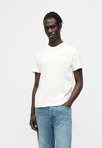 Λευκό βαμβακερό T-shirt με κοντά μανίκια και μικρό λογότυπο στο αριστερό στήθος· συνδυασμένο με ανοιχτό μπλε τζιν παντελόνι.