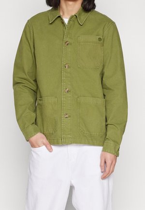 Person trägt eine grüne, geknöpfte Jacke mit drei Taschen und weiße Hose, steht vor einem einfarbigen hellen Hintergrund.