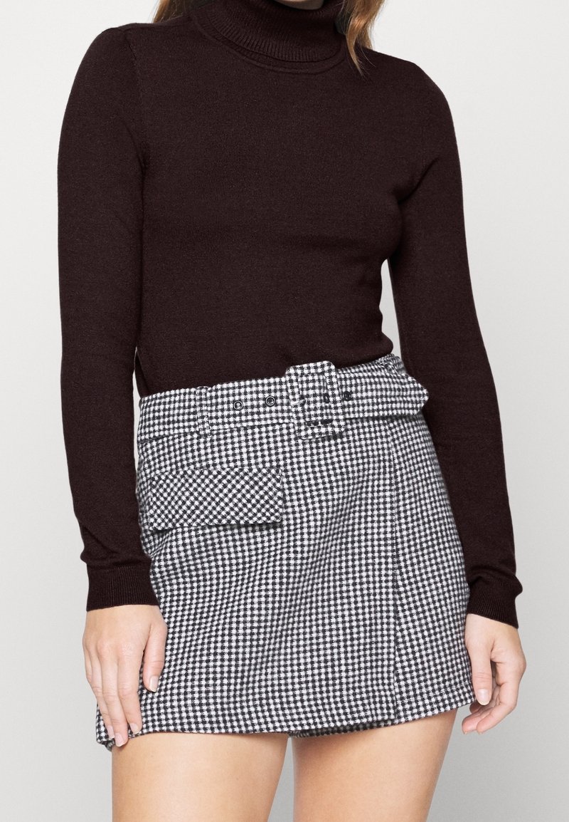Maglione a collo alto marrone abbinato a una gonna a scacchi bianca e nera con una cintura larga e un dettaglio della tasca frontale.