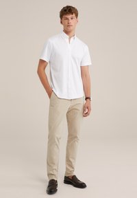 Camisa blanca de manga corta con botones, pantalones beige claro y mocasines negros. Textura suave, corte ajustado y sutil logotipo de la marca en la camisa.