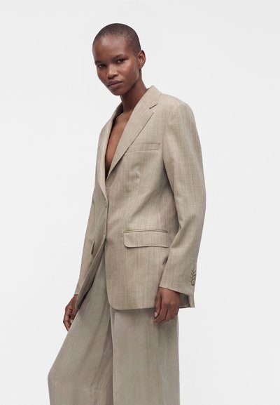 Personne portant un blazer beige oversize et un pantalon large, debout devant un fond clair uni, regardant vers la caméra.