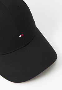 Svart baseball-cap laget av teksturert stoff. Har en buet skjerm og en liten brodert logo i rødt, hvitt og blått på forsiden.