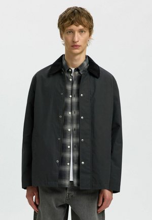 Junger Mann, der eine schwarze Jacke über einem schwarz-weißen karierten Hemd und grauen Jeans trägt und vor einem einfarbigen hellen Hintergrund steht.