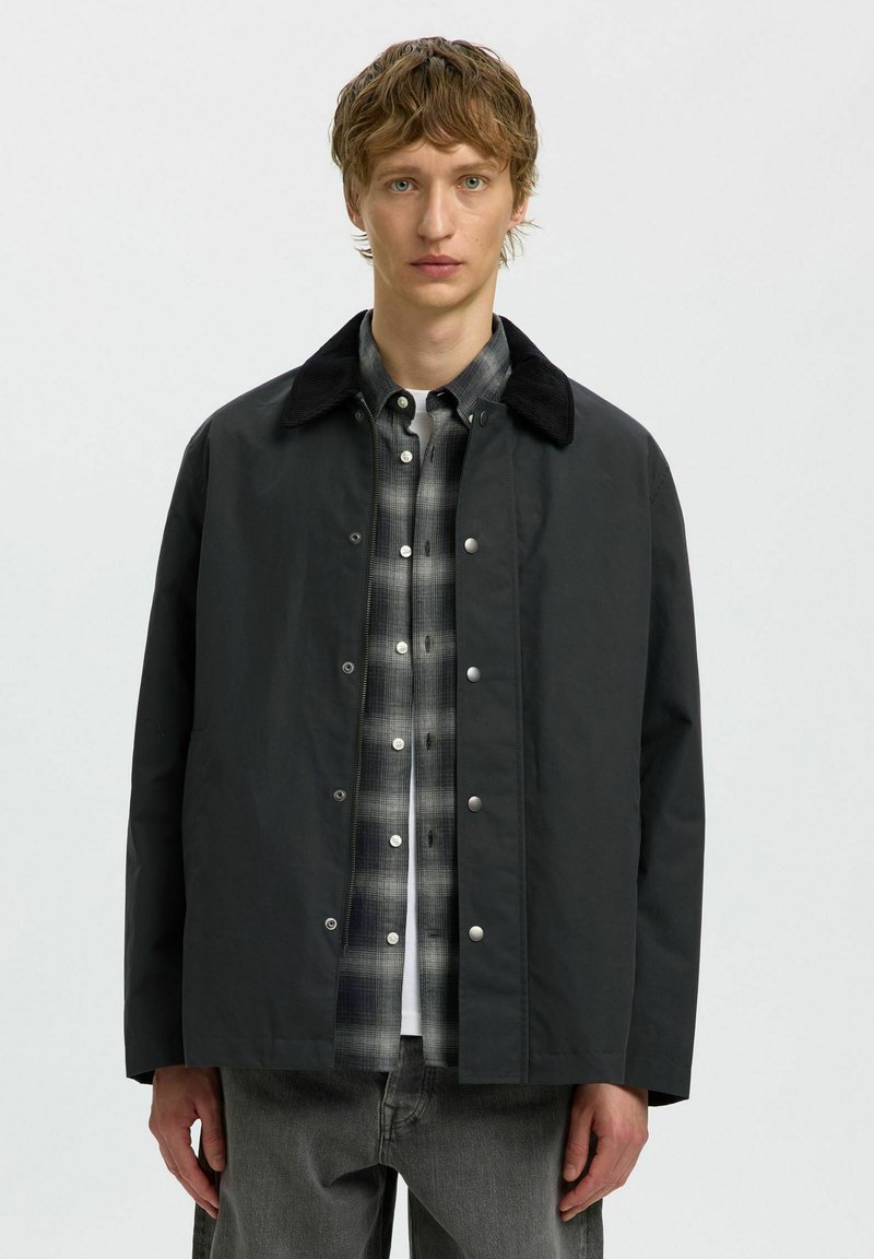 Jeune homme portant une veste noire par-dessus une chemise à carreaux noir et blanc et un jean gris, debout devant un fond clair uni.
