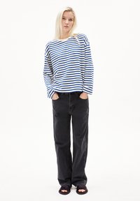 Gestreiftes Langarmhemd in Weiß und Blau; schwarze lässige Jeans; schwarze Sandalen mit offener Zehe. Model steht mit Händen in den Taschen.
