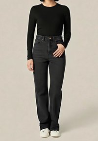 Personne portant un haut à manches longues noir, un jean foncé taille haute et des baskets blanches, debout avec une main dans une poche.