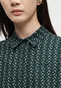 Gros plan d'une personne portant une chemise boutonnée vert foncé avec un motif de raquettes et balles de tennis, avec un petit logo de crocodile sur la poitrine.