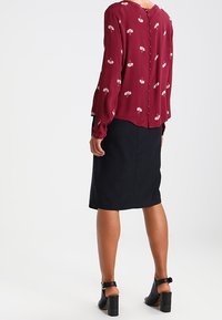 Blouse rouge à motif floral avec manches longues et fermeture par boutons au dos, associée à une jupe noire mi-longue et des sandales à talons carrés.