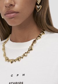 Primo piano di una persona che indossa una collana d'oro a forma di cuore e orecchini d'oro a forma di cuore abbinati, con una maglietta bianca con la scritta "CPH Studios".