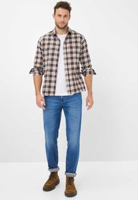 Man staat met een bruin geruit shirt over een wit T-shirt, blauwe jeans met opgerolde zomen en bruin veterschoenen op een witte achtergrond.