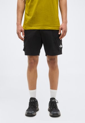 Man draagt zwarte The North Face-short, zwarte sneakers, witte sokken en een geel T-shirt, staand tegen een effen achtergrond.