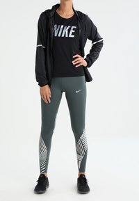 Donna in piedi, con giacca e maglietta nere Nike, leggings grigi a fantasia Nike e scarpe da ginnastica nere su uno sfondo bianco.