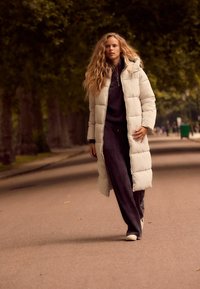 Manteau long blanc en duvet avec une capuche, associé à un pull côtelé sombre et un pantalon. Le décor présente des chemins bordés d'arbres et une route lisse.