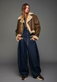 Bruine leren jas met schapenvoering, marineblauwe denim jumpsuit met grote knopen, en brede donkere jeans. Bruine sneakers maken de outfit compleet.