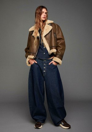 Braune Lederjacke mit Schaffellfutter, marineblaue Denim-Jumpsuit mit großen Knöpfen und weit geschnittene dunkelblaue Jeans. Braune Sneaker runden das Outfit ab.