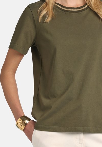 Femme portant une chemise à manches courtes vert olive avec un col rayé et un bracelet manchette doré, main dans la poche d'un pantalon beige.