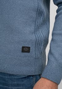 Pulover albastru dintr-un material moale, texturat, cu detalii în dungi pe laturi și o etichetă neagră cu marcaj aproape de tiv.