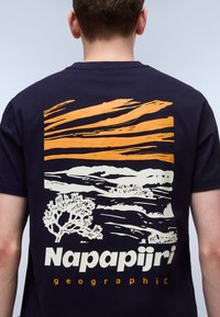 Camiseta de algodón azul marino con un gráfico en la parte posterior de líneas horizontales naranjas y cremosas con un diseño de paisaje y árbol. Nombre de la marca impreso debajo.