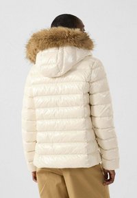 Giacca puffer crema con finitura lucida, design a cucitura orizzontale e cappuccio foderato in pelliccia marrone. Vestibilità aderente con polsini elasticizzati.