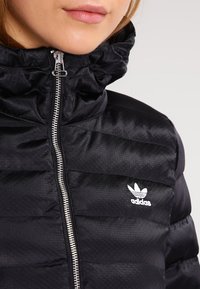 Svart puffjacka med hög krage, med blixtlås fram och vitt Adidas-logotyp på vänstra bröstet. Slät, texturerad tyg.