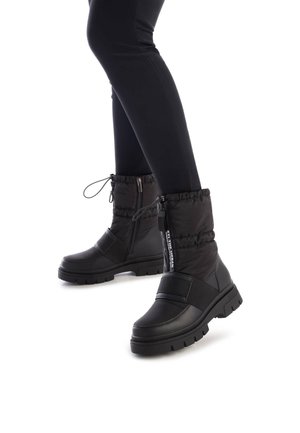 Botas negras hasta el tobillo con parte superior texturizada y acolchada, que cuentan con un cierre lateral y una correa elástica. Suela de goma gruesa con un patrón de relieve.
